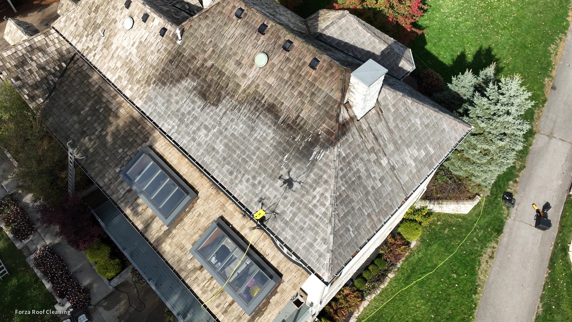 4K drone still showing clear cleaning line on cedar shake roof - left side treated and lighter, right side untreated and dark, washing drone hovering over Muirfield Village home
