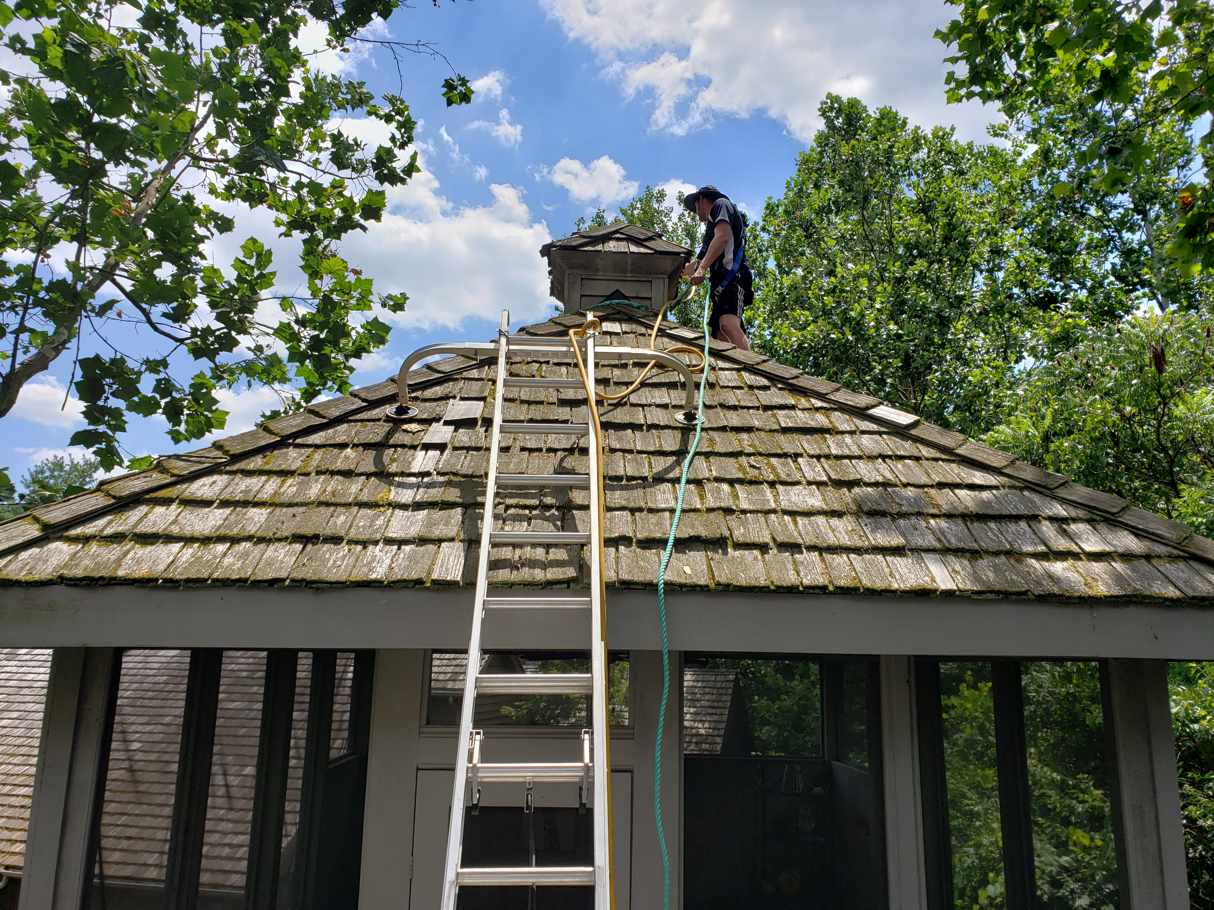 Forza technician wearing safety harness on elevated porch roof near cupola, ladder visible, green safety rope, cedar shake cleaning, The Quarry Dublin Ohio