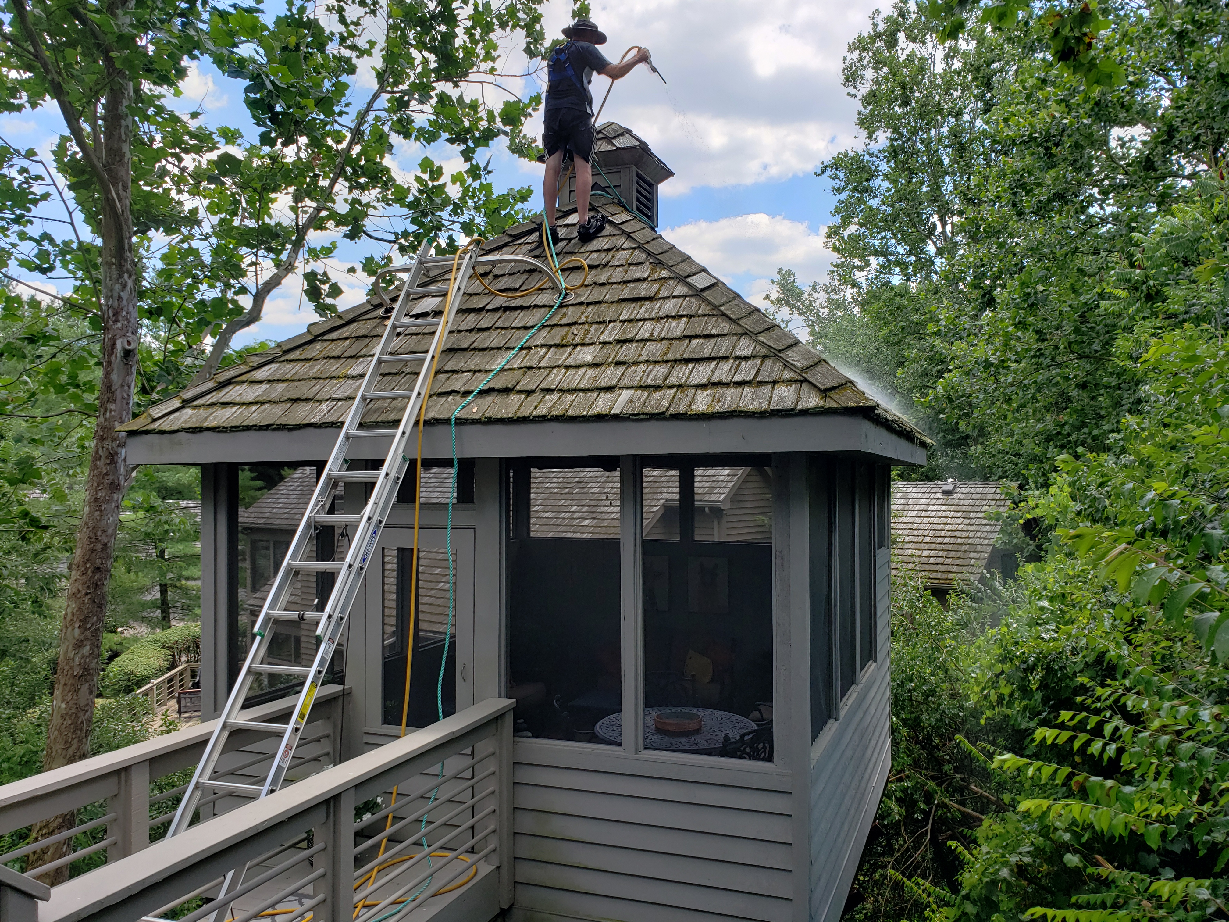 Forza technician with safety harness spraying treatment on elevated porch roof, walkway visible below, cedar shake cleaning, The Quarry Dublin Ohio