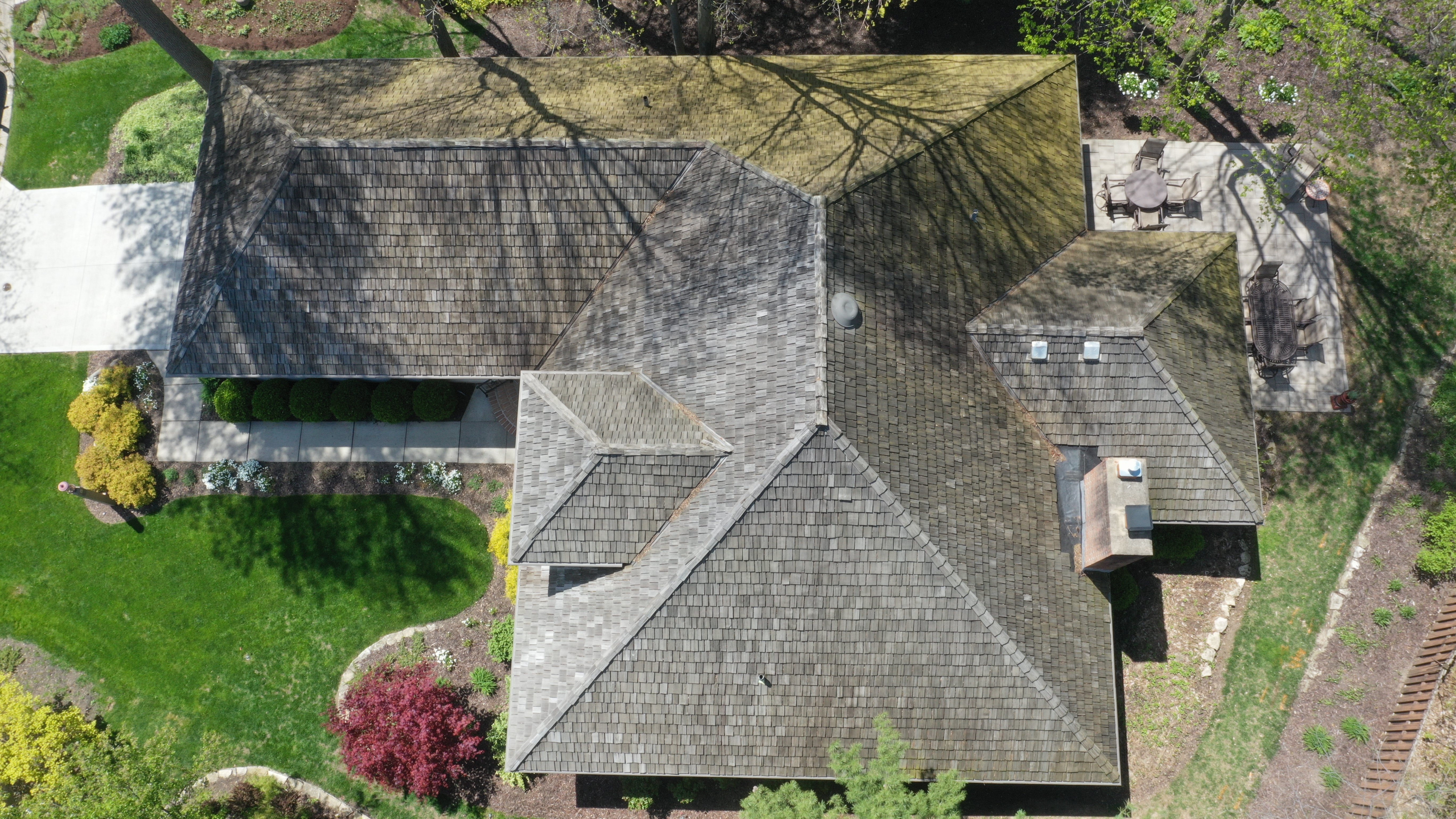 Before cedar roof cleaning - overhead view of cedar shake roof with gray weathering and heavy green algae on rear slopes near tree canopy, white siding home with covered porch, The Sanctuary Columbus Ohio