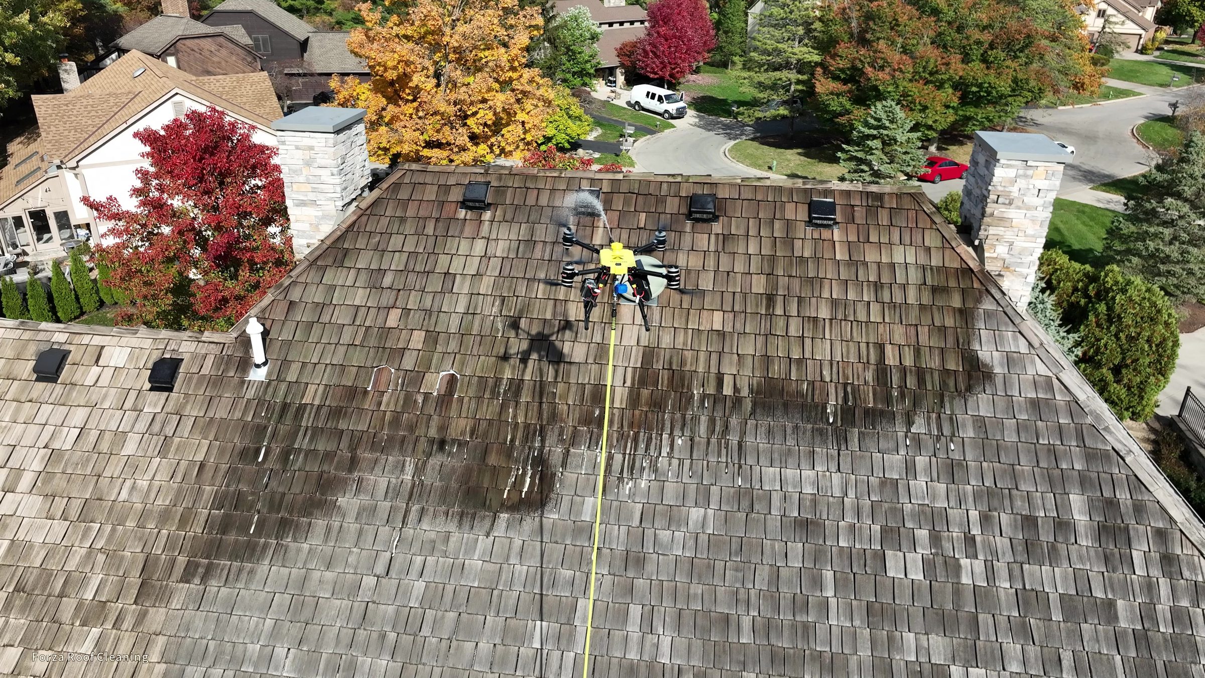 4K drone still showing Forza washing drone spraying front of cedar shake roof in Muirfield Village, spray mist visible, fall foliage neighborhood